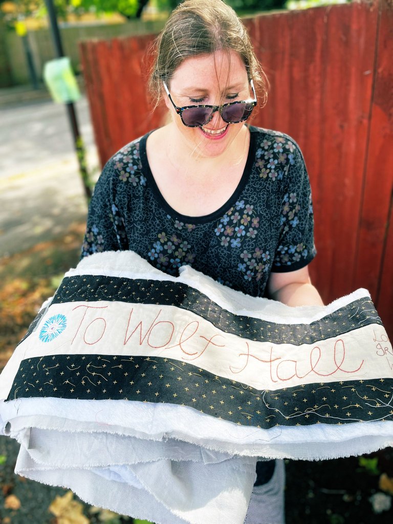 A woman in dark glasses and a black dress holds a pile of quilted fabric. The words To Wolf Hall and a cornflower are visible in the stitching.