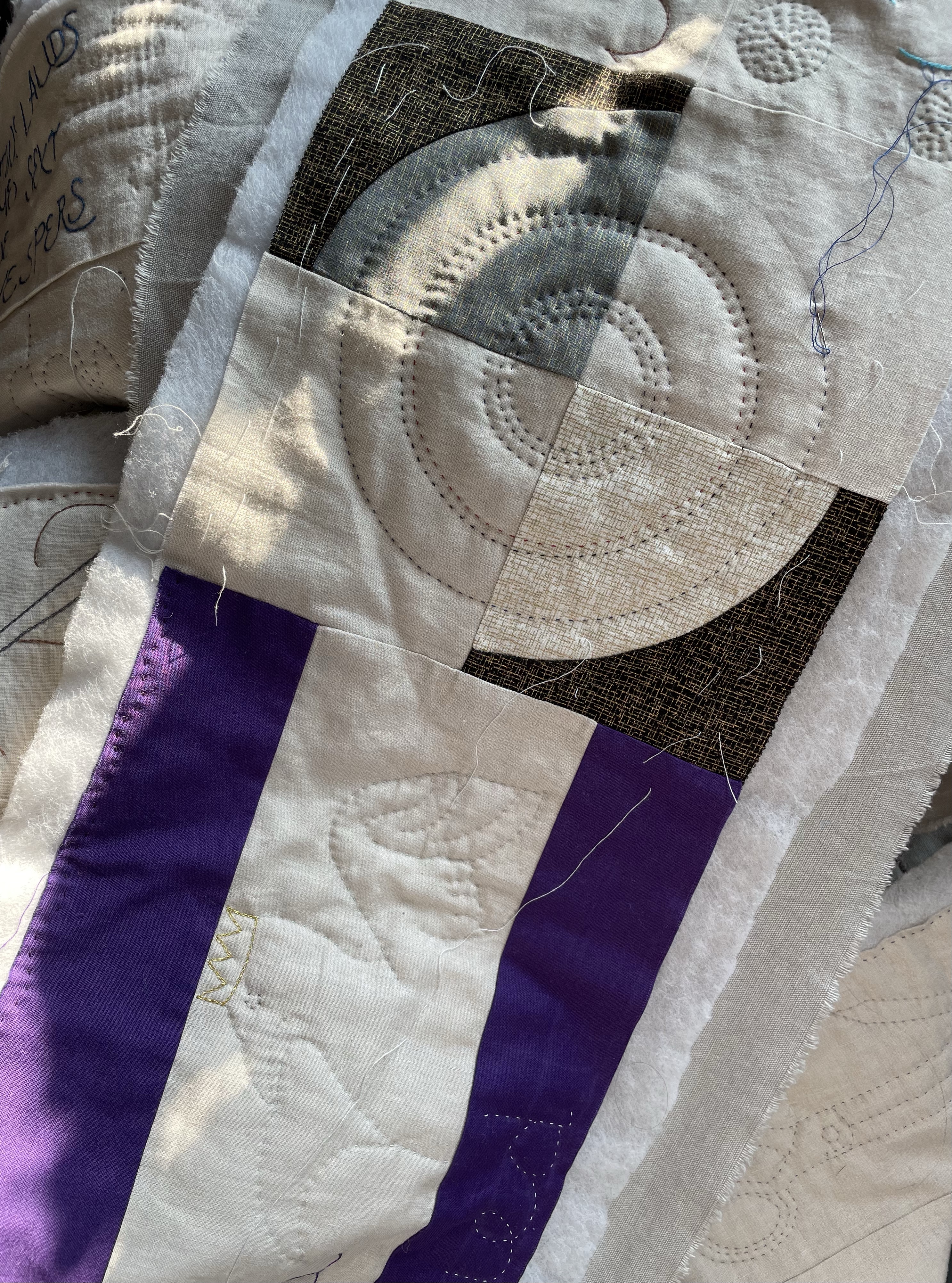 Quilted fabric strips, in cream, purple, black, and grey. A quilted falcon is visible, and a curved quilt block is being quilted with circles, a needle and thread are waiting to be stitched in.
