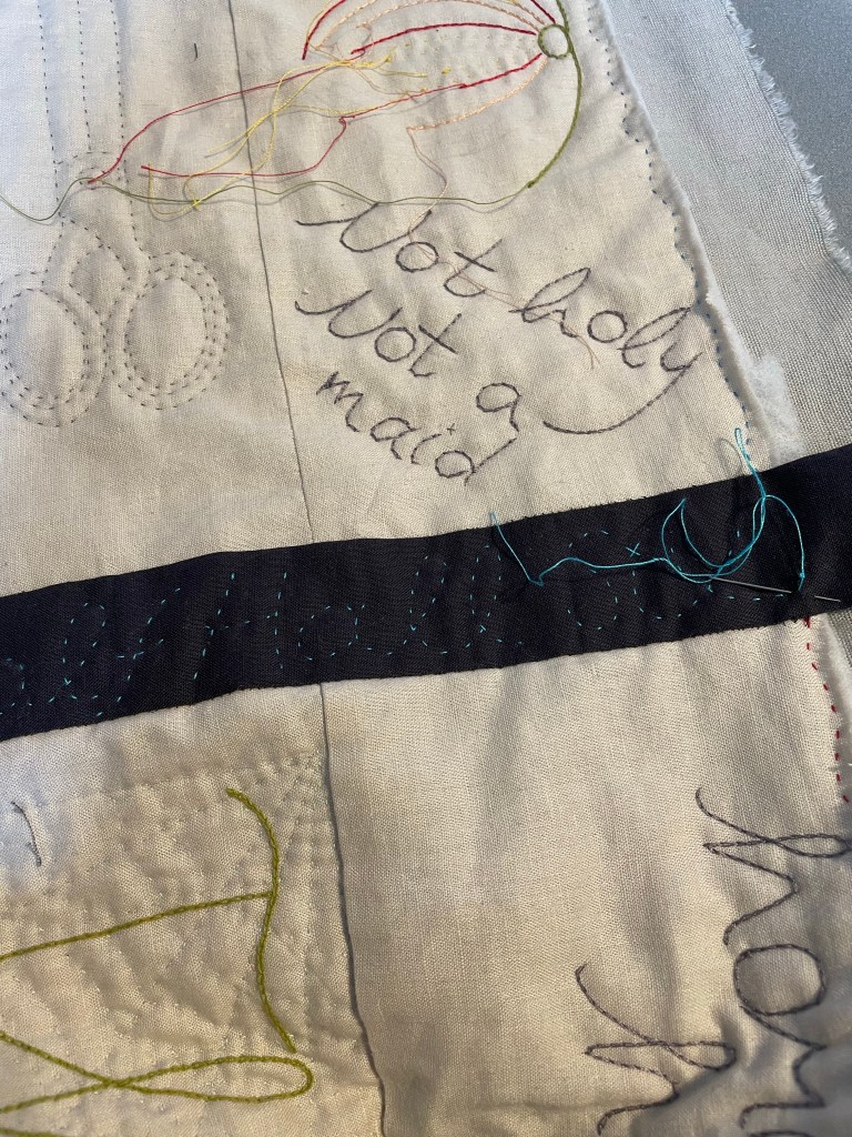 Pieces of quilted fabric in cream with decoration; a dark grey strip joining two pieces with blue text stitching and a needle and thread ready to set a stitch.