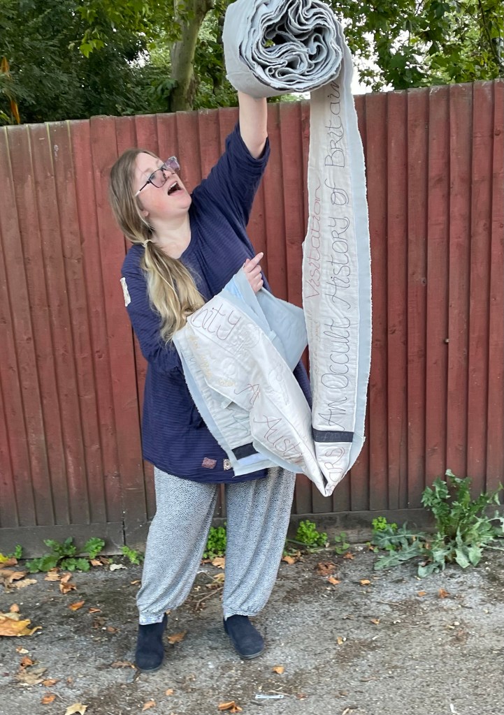A woman in a blue dress and grey trousers holds up a long pieces of quilted work against a backdrop of a brown fence. 