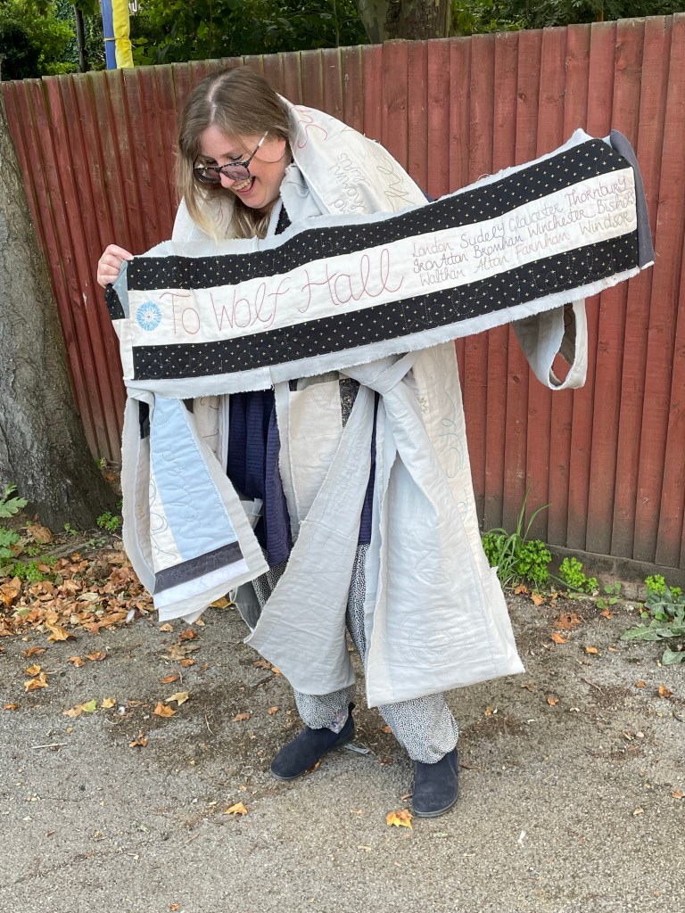 A woman in a blue dress and grey trousers holds up a long pieces of quilted work against a backdrop of a brown fence. The quilted lettering reads “To Wolf Hall”