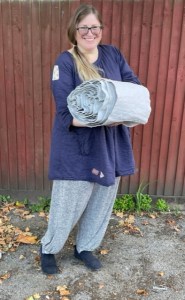 A white woman stands in front of a fence, holding a rolled piece of quilting which is predominantly pale grey.