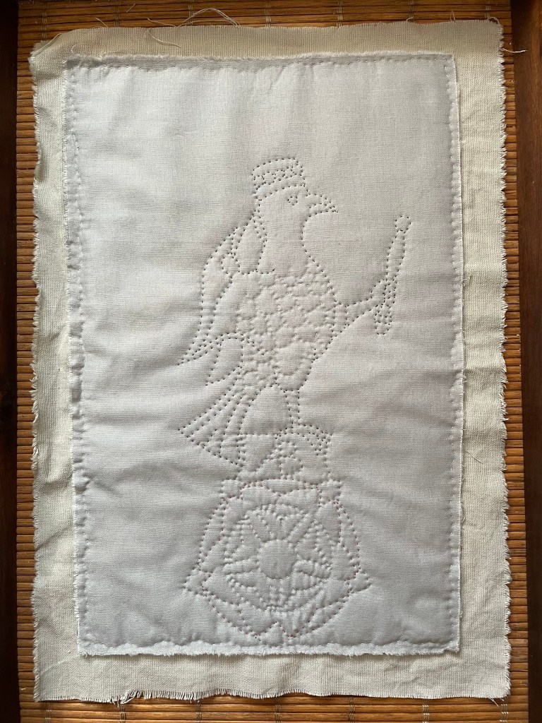 A grey quilted piece of a falcon, which is standing on top of a Tudor rose. The falcon is crowned and holds a sceptre in its claw.