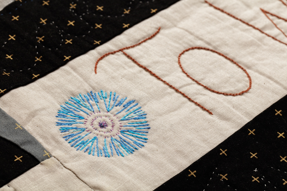 A quilted cornflower, and the word 'To' stitched onto cream cloth, black cloth borders with gold crosses.