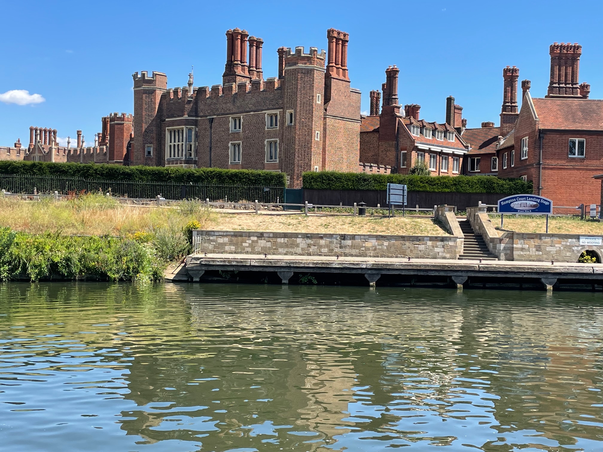 Hampton Court Palace - a brick building with numerous tall chimneys is shown on the opposite bank of the river on a sunny day. The Palace is reflected in the ripples of the water.