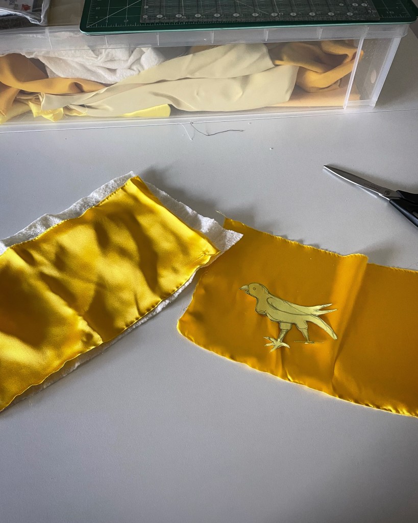 Pieces of yellow silk, laid on a white surface. A yellow cardboard template of a bird rests on one piece of silk. A pair of scissors is just visible. 