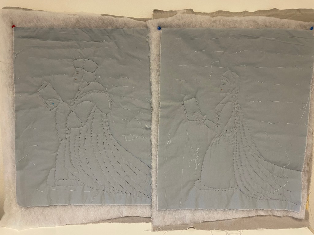 Two quilted figures on grey fabric: one a man wearing long robes and a hat; the woman wearing a long dress and cloak. Both are kneeling and holding books