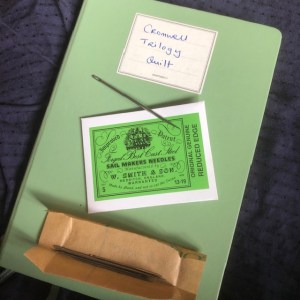 A green notebook with a packet of sail makers' needles resting on it. The needles are out of the packet and can be seen to be large and thick,