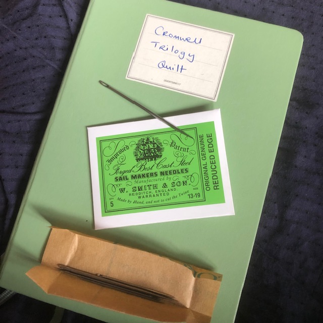 A green notebook with a packet of sail makers' needles resting on it, one of the needles is out of the packet and is large.