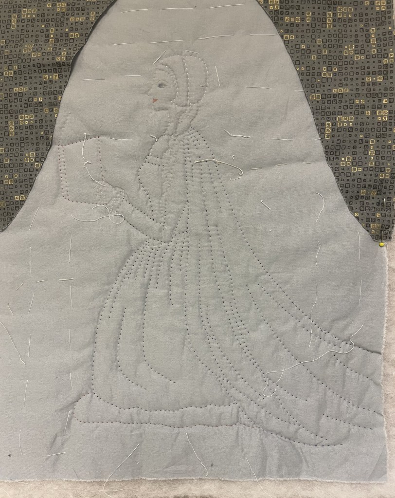 A hand quilted figure of a kneeling woman, wearing a cloak, long skirts, and holding a book.